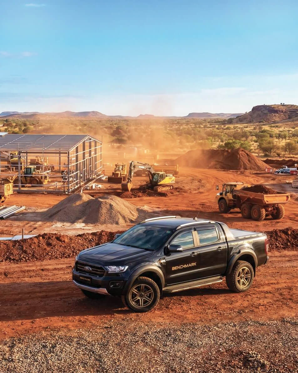 Black pickup truck with Benchmark branding parked on a red dirt construction site with heavy machinery and steel framework building in the background under a clear blue sky.