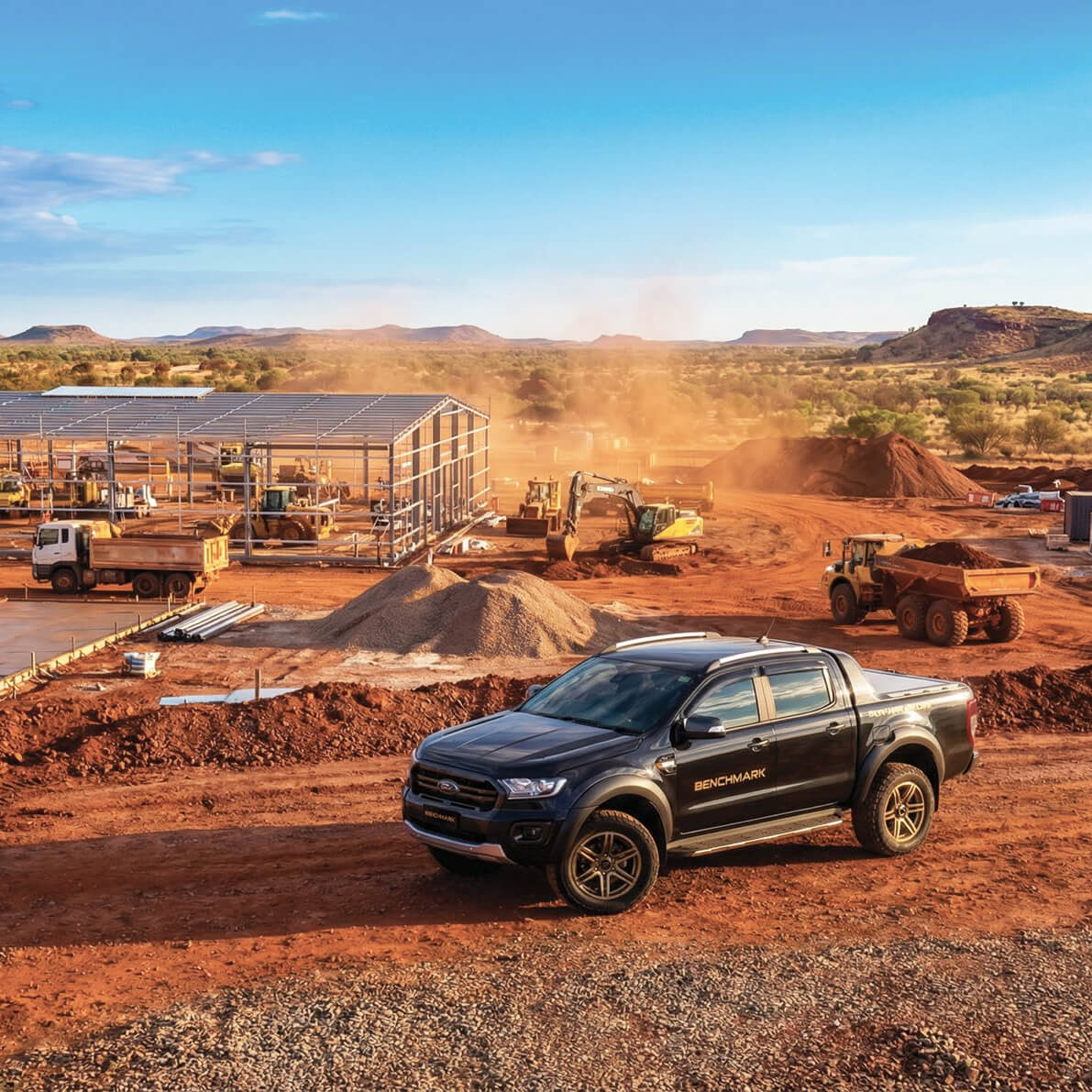 Black pickup truck with Benchmark branding parked on a red dirt construction site with heavy machinery and steel framework building in the background under a clear blue sky.