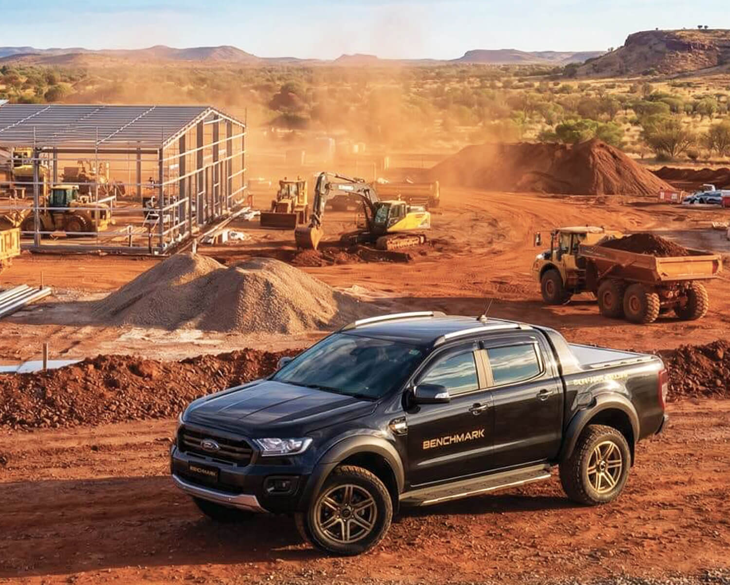 Black pickup truck with Benchmark branding parked on a red dirt construction site with heavy machinery and steel framework building in the background under a clear blue sky.