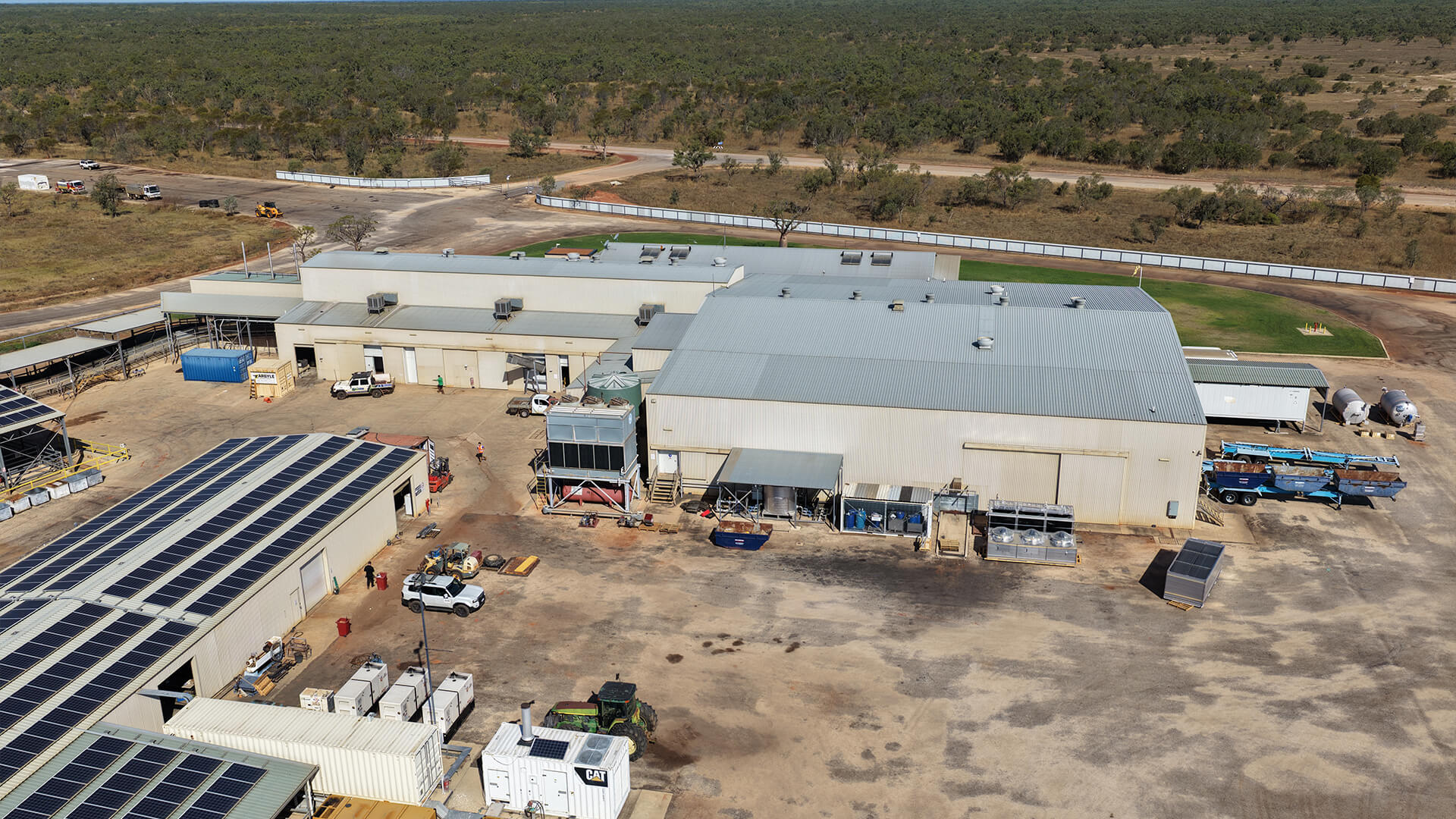 Aerial view of an industrial facility with solar panels on roofs and vehicles on a dirt lot.