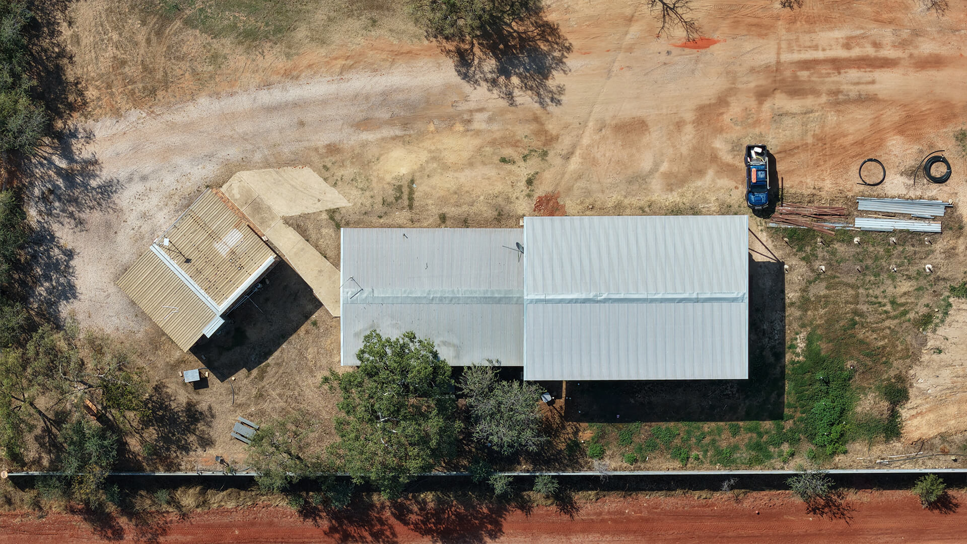 Aerial view of a rural property with two buildings, scattered trees, dirt road, and a parked blue vehicle.