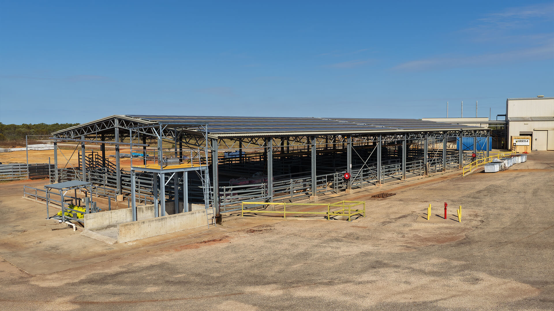 Large metal livestock pen with roof, surrounded by dry ground and a concrete platform under clear sky.