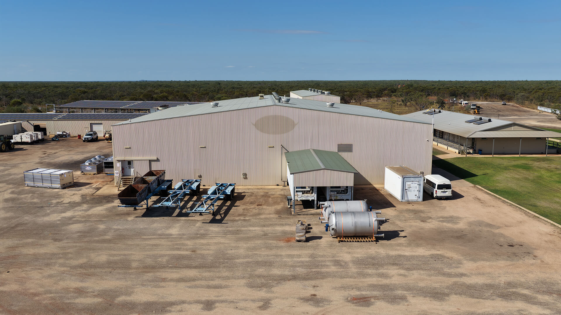 A large industrial building with outdoor machinery, vehicles, and storage tanks on a dirt lot.